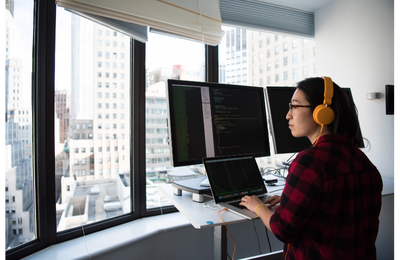 A woman is working at a laptop with two large monitors at a desk in a high-rise building with large windows.