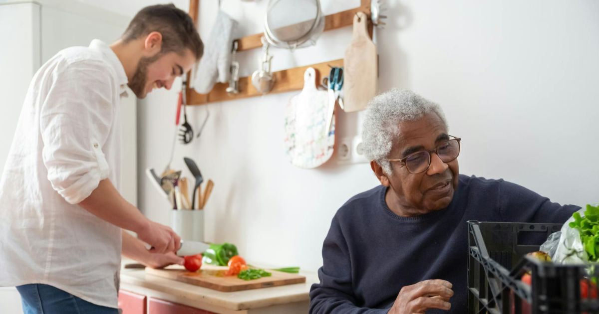 Man cutting vegetables on a counter while an elderly man looks at groceries on a table.